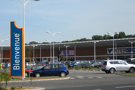 photo du parking avec des voitures et des magasins en fond
